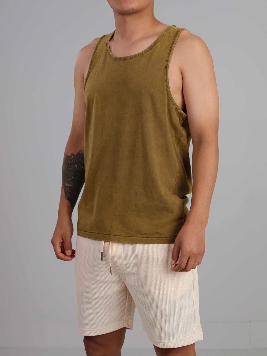 Against a plain gray background, someone wears a Settler Snow Vintage Wash Tank Top by ID Menswear with beige shorts. The lightweight brown sleeveless shirt highlights the tattoo on their left arm, adding an edge to the classic ensemble.