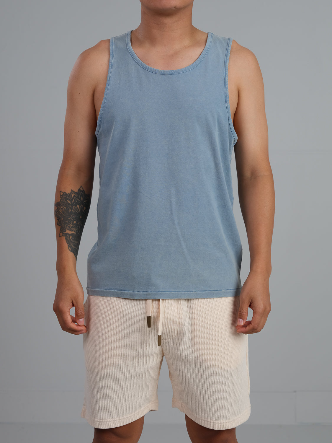 Against a gray backdrop, someone showcases a tattoo on their left arm while wearing the ID Menswear Settler Snow Vintage Wash Tank Top and beige shorts.