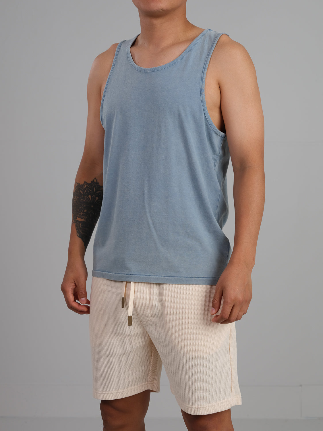 Dressed in ID Menswears Settler Snow Vintage Wash Tank Top and light blue, cream-colored organic cotton shorts, a person stands against a plain gray background, exuding a vintage vibe with a tattoo on their left arm.