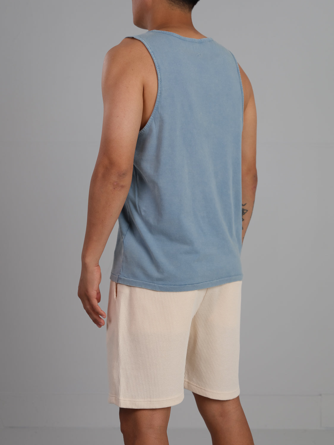 A person in an ID Menswear Settler Snow Vintage Wash Tank Top and beige shorts stands against a plain gray background, evoking a vintage look as they face away.