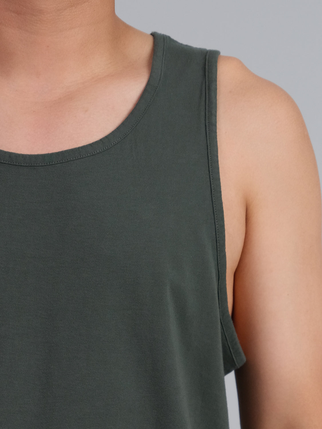 A closeup shot captures a person in an ID Menswear Settler Organic Cotton Tank Top. The dark green top, showcased against a light blue background, highlights its texture and color from chest to shoulder.