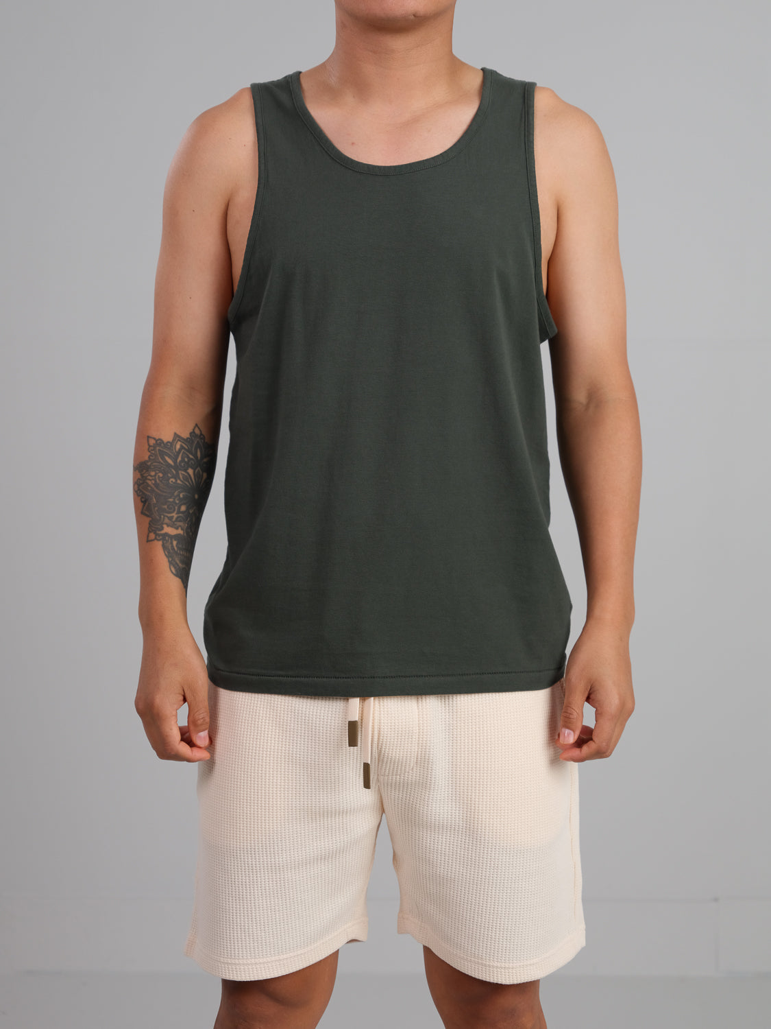 A person models a dark green Settler Organic Cotton Tank Top from ID Menswear, paired with light-colored shorts against a plain gray background. The lightweight, regular-fit organic cotton tank top subtly reveals a tattoo on their left upper arm while their face remains out of view.