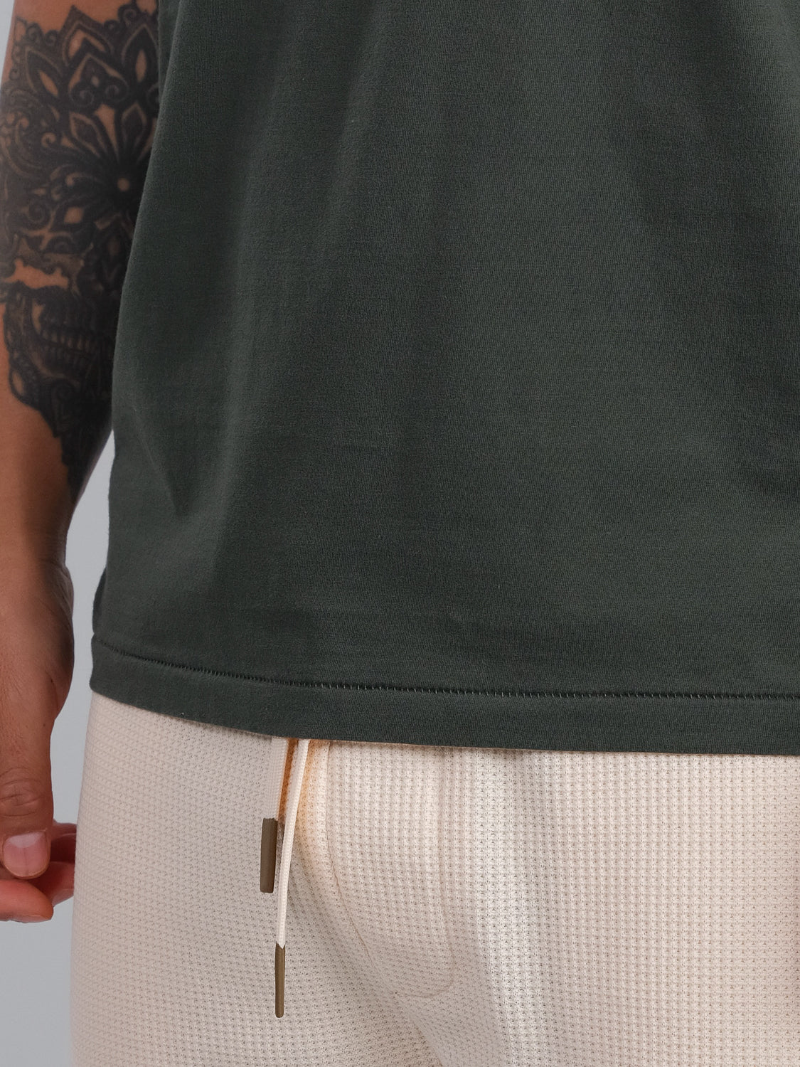 Close-up of someone wearing an ID Menswear dark green Settler Organic Cotton Tank Top and light beige textured drawstring pants. A detailed tattoo decorates their arm while their hand rests relaxed at their side.