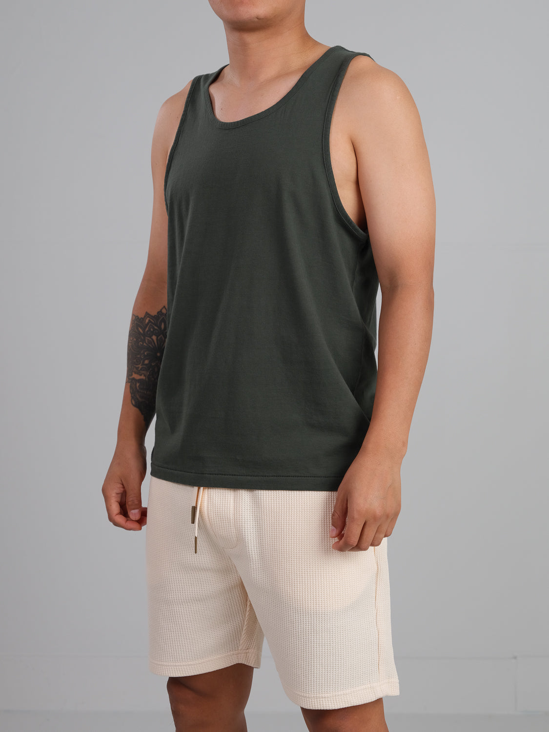 A person wears the Settler Organic Cotton Tank Top from ID Menswear, a dark green, lightweight top made of organic cotton, paired with light beige shorts. They showcase a tattoo on their left arm against a plain gray background.