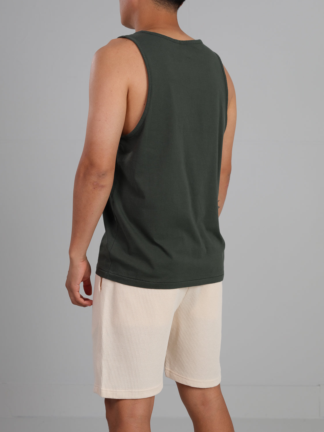 Facing away, a person wears ID Menswears lightweight Settler Organic Cotton Tank Top in dark green and light beige shorts against a plain light gray background.