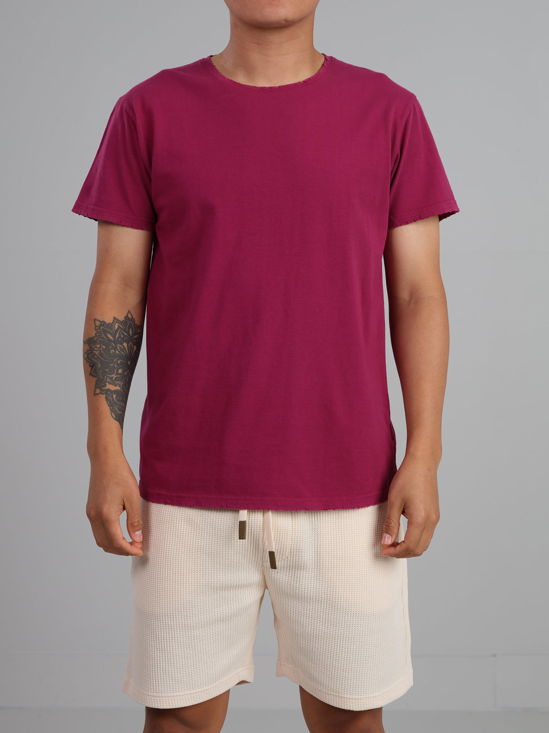 A person stands against a gray background, wearing a maroon Trekker Tattered Crew Neck Tee by ID Menswear and beige shorts. The geometric tattoo on their left arm complements the casual look, while the lightweight jersey tee provides comfort and highlights their torso.