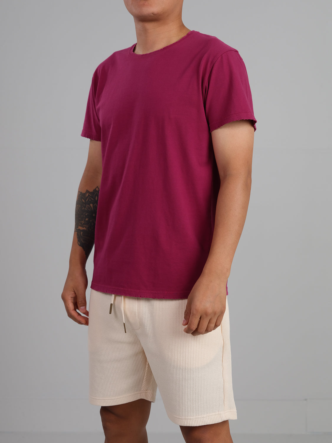 Against a gray backdrop, an individual wears a maroon Trekker Tattered Crew Neck Tee by ID Menswear with cream shorts. A tattoo on their right forearm is visible as they confidently pose, left hand on hip.