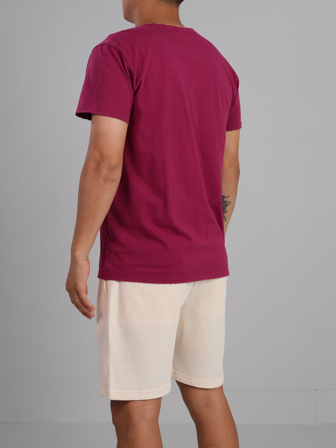 A person is seen from behind, wearing the ID Menswears Trekker Tattered Crew Neck Tee in organic cotton with beige shorts. A small tattoo is visible beneath the short sleeve, contrasting subtly against a plain gray wall.