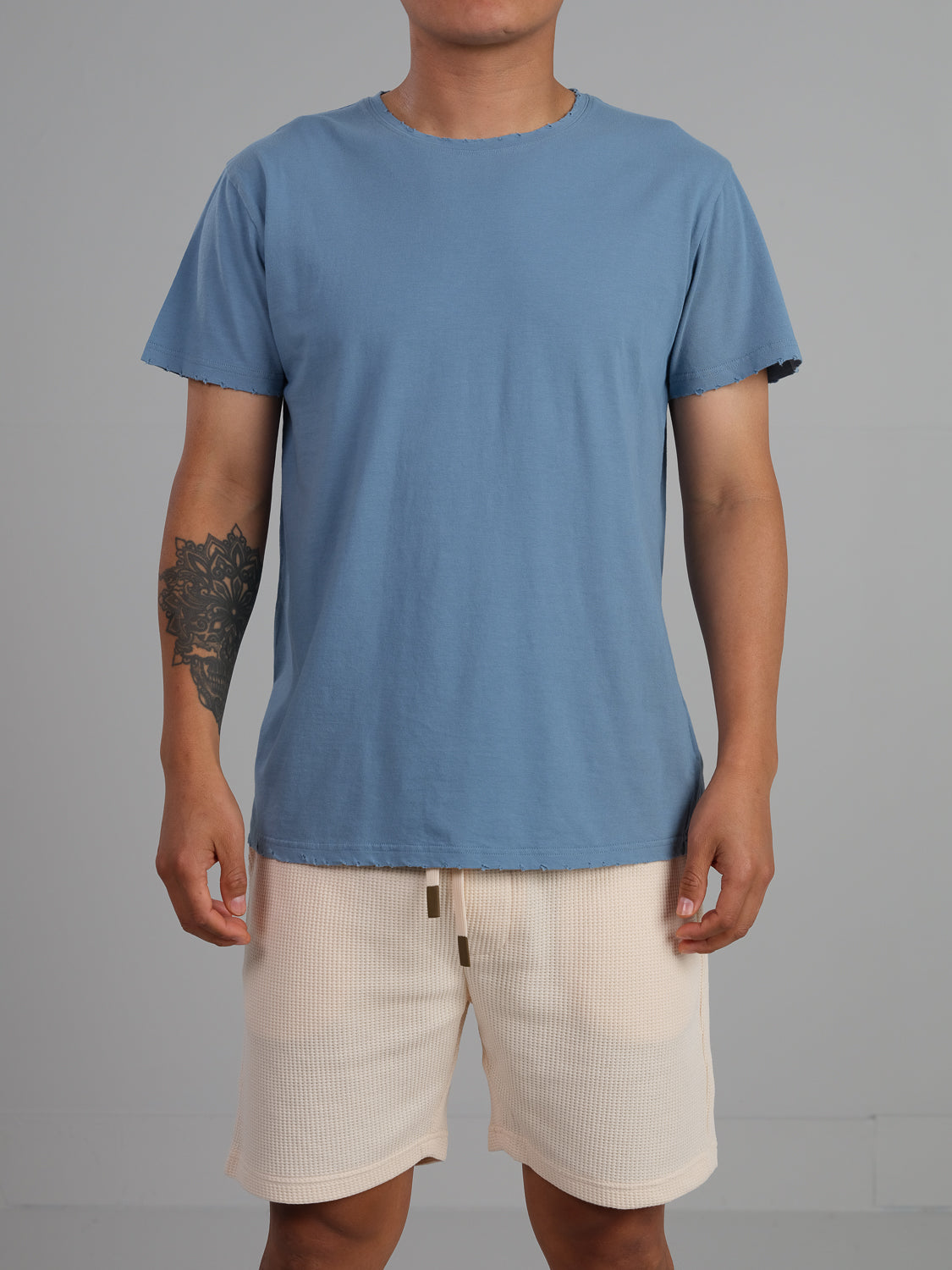 Against a gray backdrop, a person wears a light blue lightweight jersey and beige shorts. Their right arm sports a tattoo. The casual outfit is completed with an organic cotton Trekker Tattered Crew Neck Tee by ID Menswear, and they face forward confidently.