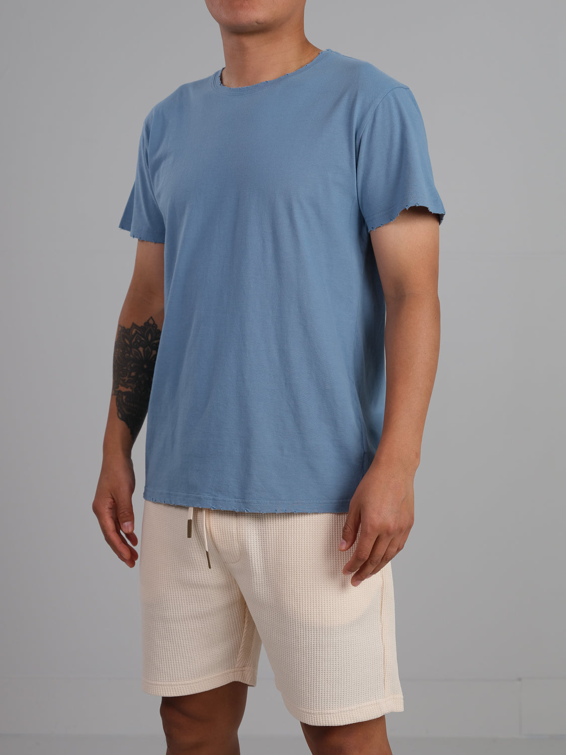 A person in a lightweight ID Menswear Trekker Tattered Crew Neck Tee stands against a gray background, wearing beige shorts. A tattoo is visible on their left forearm.
