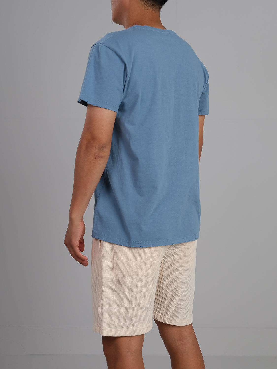 A person stands against a gray background, facing away, wearing a lightweight Trekker Tattered Crew Neck Tee by ID Menswear in blue, paired with beige shorts.