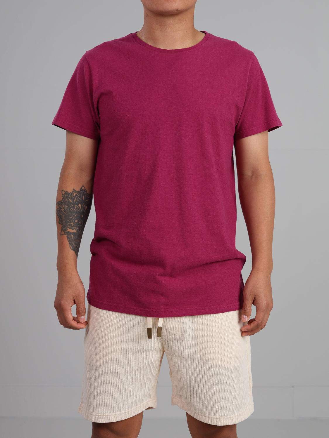 A person is wearing the Dope Hemp Organic Cotton Tee by ID Menswear in maroon with light beige shorts. Their arms are down by their sides, showing a tattoo on one forearm against a plain gray background.