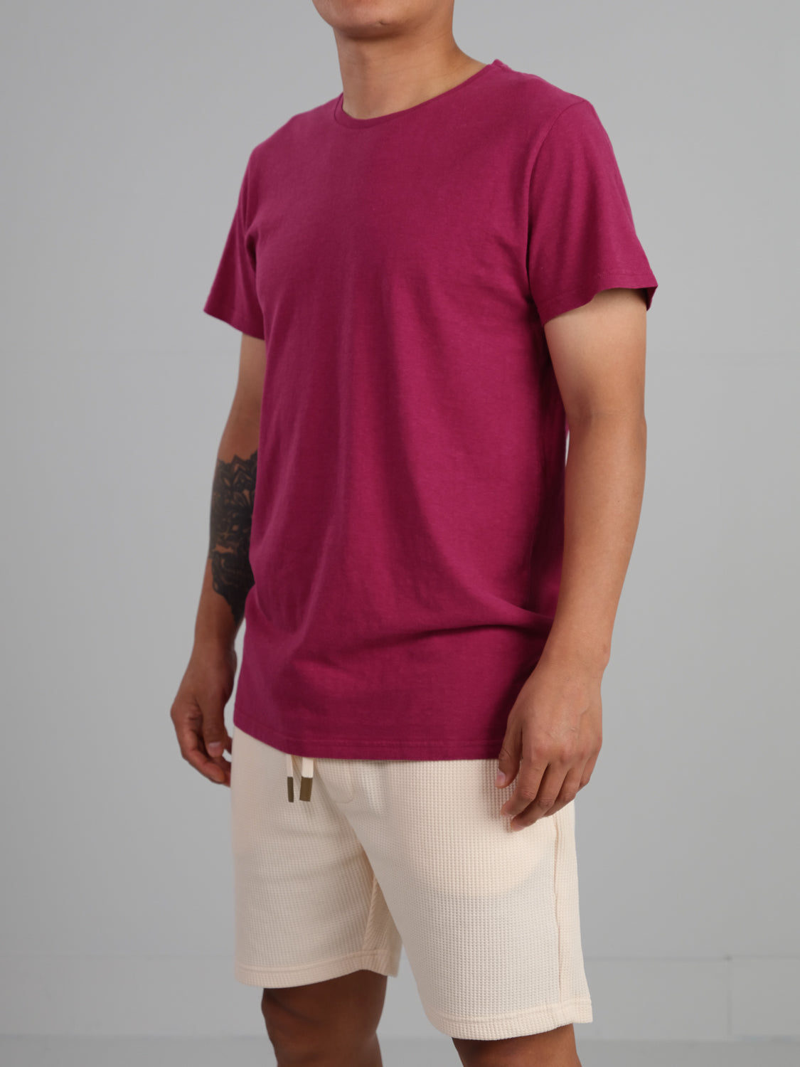 Against a plain backdrop, an individual wearing the Dope Hemp Organic Cotton Tee by ID Menswear stands with beige shorts. A tattoo graces their left forearm, and the focus is on their upper body and eco-friendly attire.