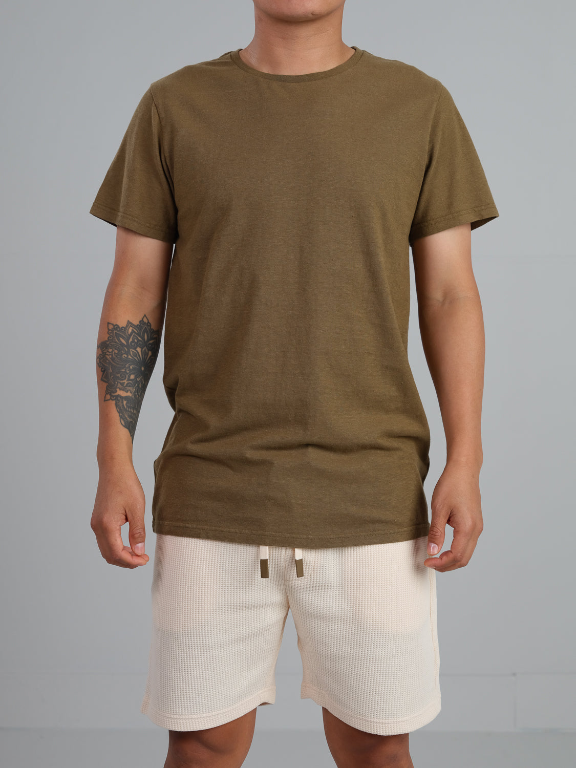 A person wearing an ID Menswear Dope Hemp Organic Cotton Tee in olive green and cream shorts stands against a plain gray background, with a tattoo visible on their arm.