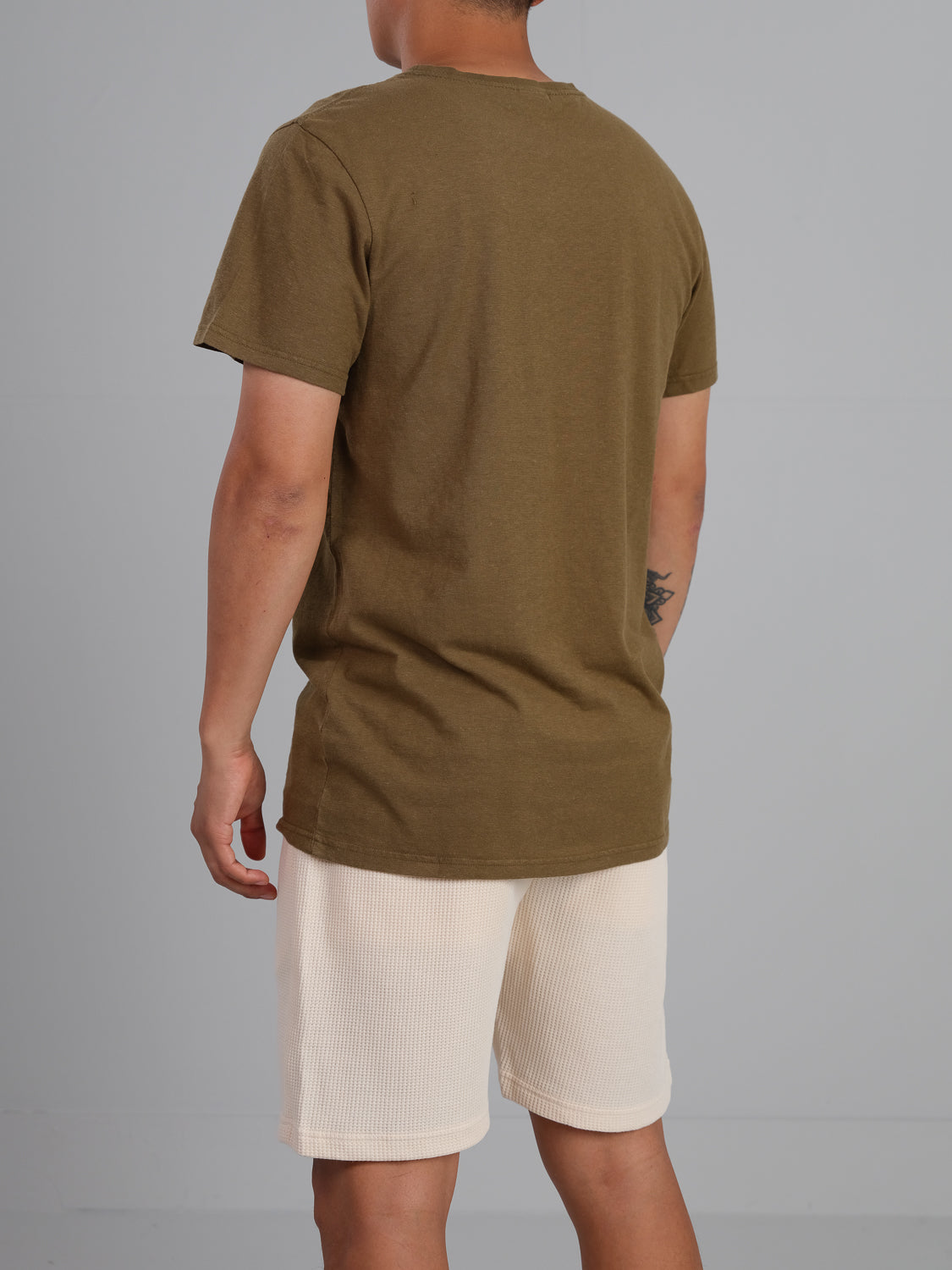 A person is seen from the back wearing ID Menswears Dope Hemp Organic Cotton Tee in olive green paired with light beige shorts. They have a small tattoo on their left forearm, set against a plain gray backdrop.