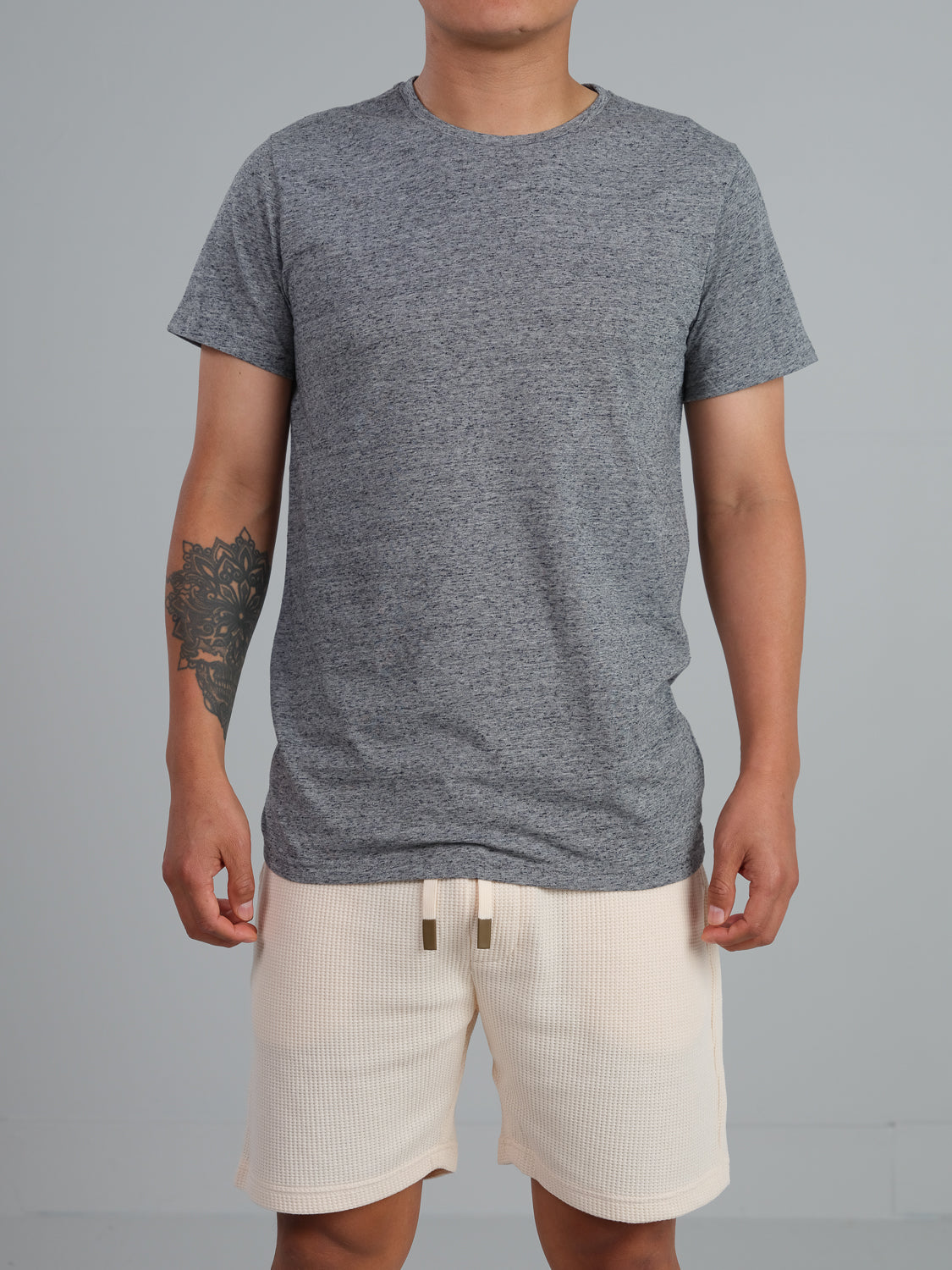 Wearing an Outclass Speckled Crew Neck Tee by ID Menswear made of cotton/poly jersey and light shorts, a person stands against a plain backdrop with a tattoo on their left forearm.