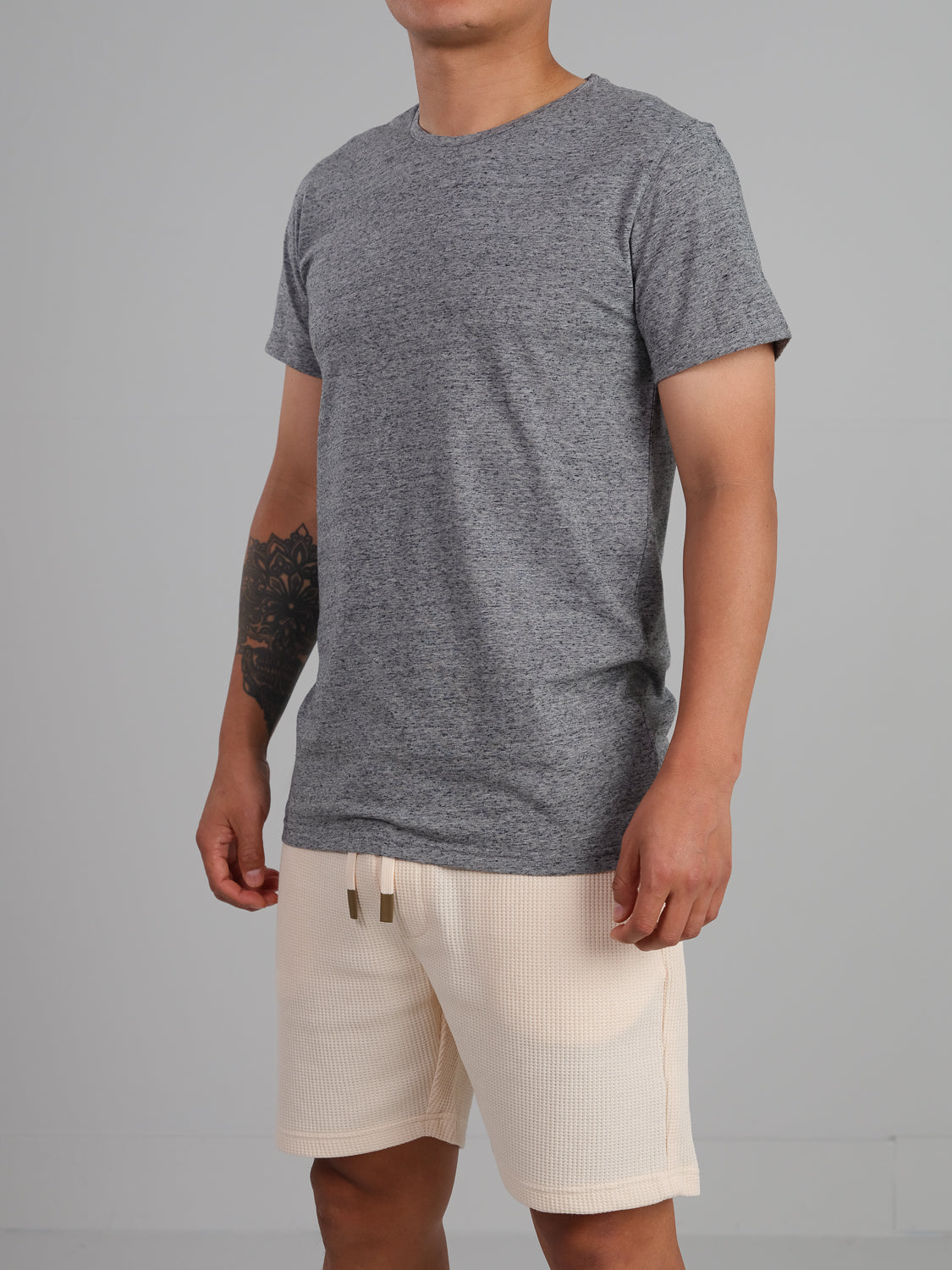 Wearing the Outclass Speckled Crew Neck Tee by ID Menswear and light-colored shorts, a person stands with arms at their sides against a plain background. A tattoo on their right forearm adds a unique touch to this everyday ensemble.