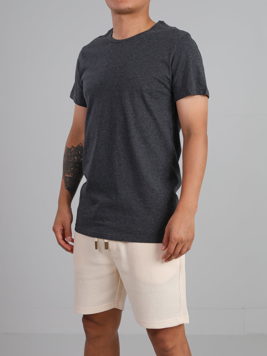 A person in an Outclass Cotton Crew Neck Tee from ID Menswear, dark gray and lightweight cotton jersey, stands in light beige shorts. Facing slightly to the side, a tattoo on their left forearm is visible against a plain gray backdrop.