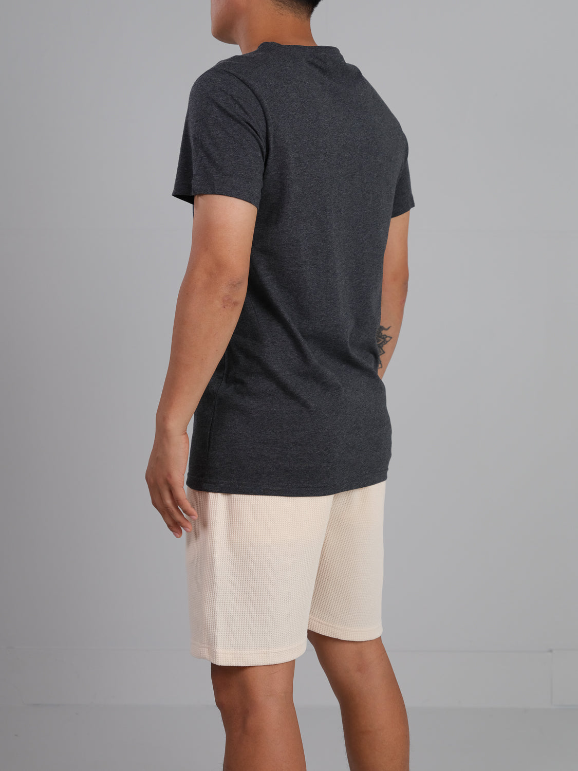 Facing away, the person wears an ID Menswear Outclass Cotton Crew Neck Tee in dark grey paired with light-colored shorts against a plain, light grey background.