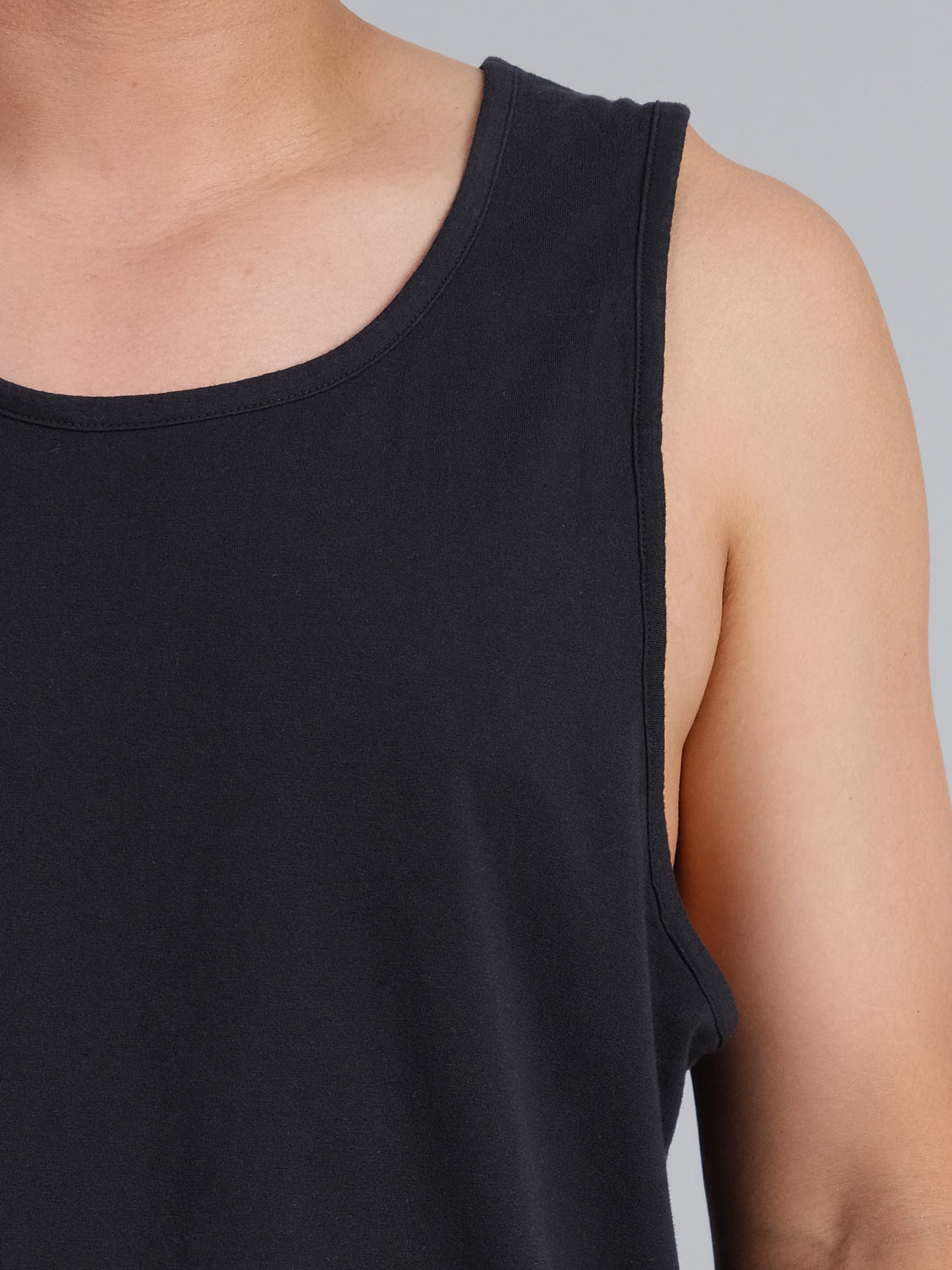 Close-up of a shoulder and upper arm wearing ID Menswears Settler Organic Cotton Tank Top. The black fabric texture and natural skin tone stand out against a gray backdrop.