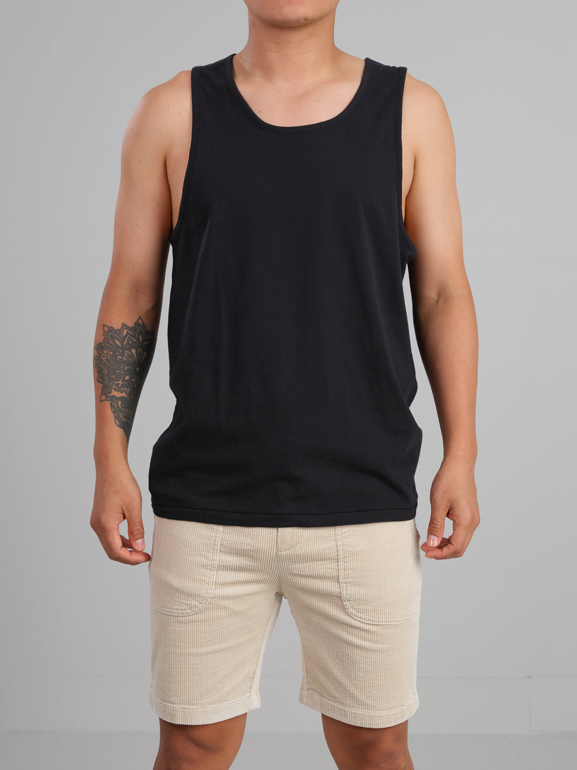 A person wears an ID Menswear Settler Organic Cotton Tank Top, showcasing a regular fit in black, paired with beige shorts. They stand against a gray backdrop, displaying a tattoo on their right arm.