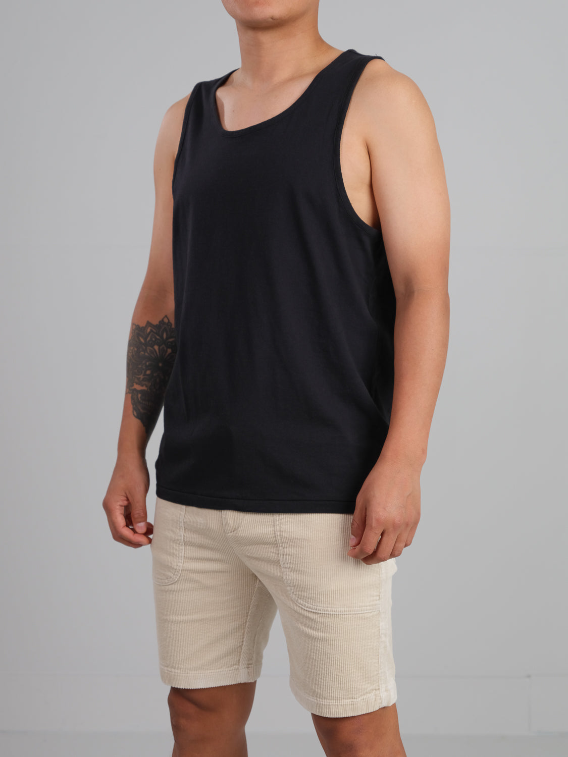A person in an ID Menswear Settler Organic Cotton Tank Top and beige shorts stands against a plain gray background, showing off a tattoo on their right arm.