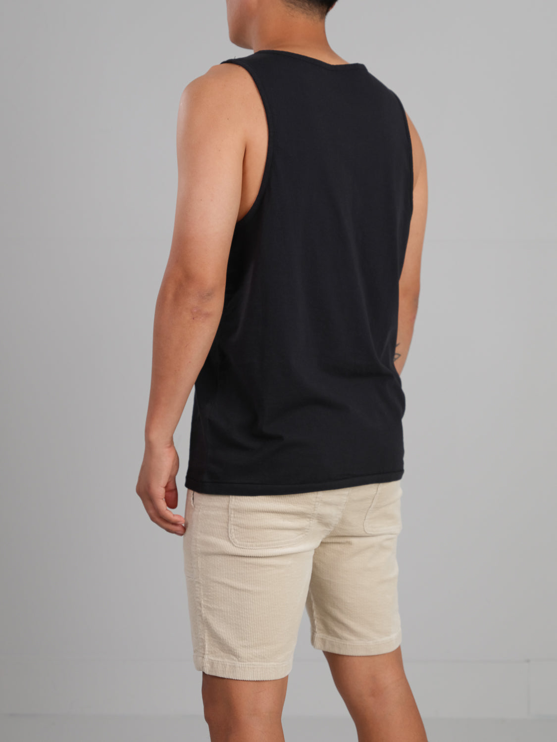 A person stands with their back to the camera against a gray background, wearing a black Settler Organic Cotton Tank Top by ID Menswear and beige shorts. Their arms hang relaxed, highlighting the tanks regular fit.