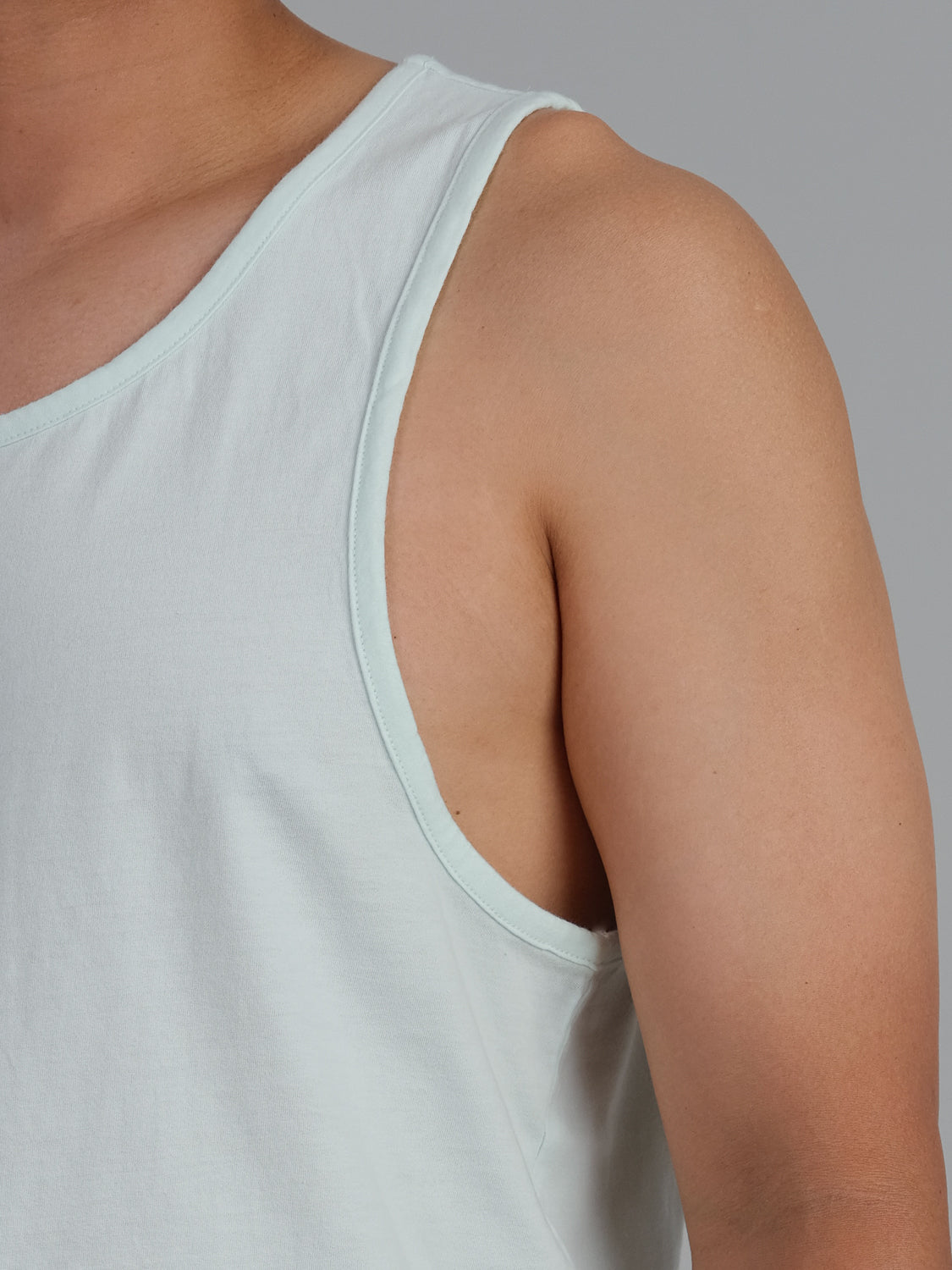 A person with medium skin tone is shown from shoulder to waist wearing a Settler Organic Cotton Tank Top by ID Menswear. The light blue, lightweight tank made of organic cotton fits perfectly. Half the upper body is visible against a gray background.