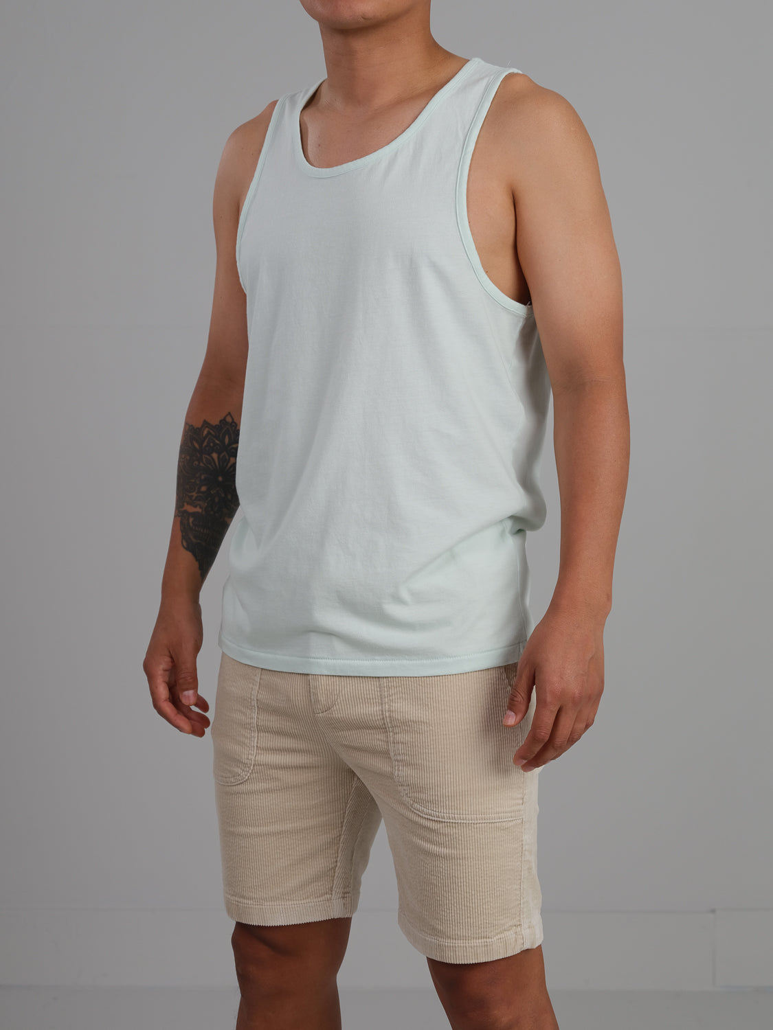 A person wears the Settler Organic Cotton Tank Top by ID Menswear in light blue with beige shorts, revealing a right arm tattoo against a plain gray background.