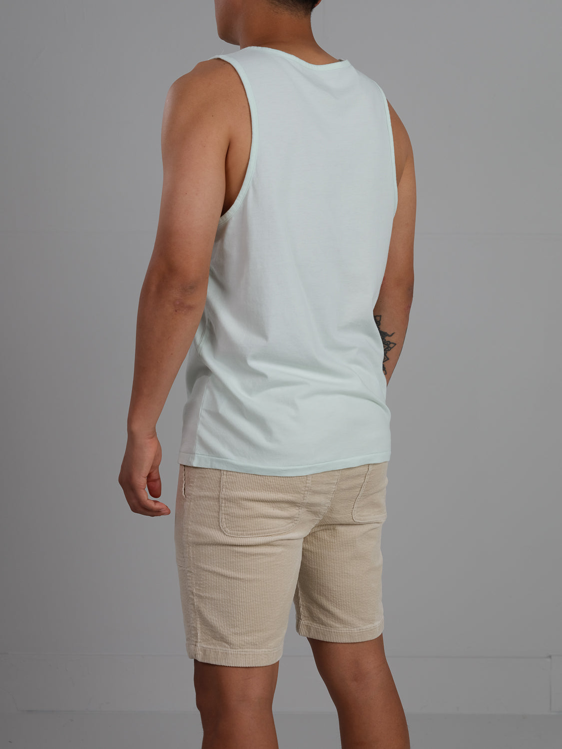 In front of a plain gray background, a person stands with their back to the camera, wearing ID Menswears Settler Organic Cotton Tank Top in light blue paired with beige shorts.