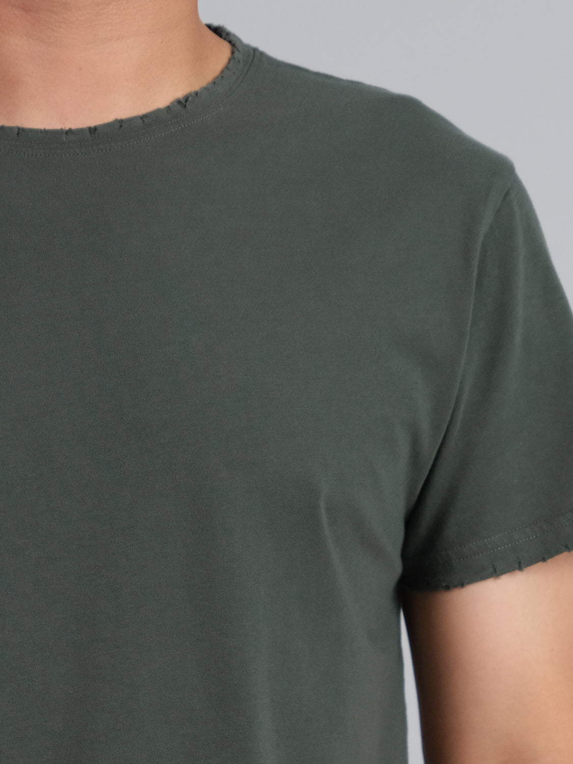 A close-up displays a person wearing a plain, dark green Trekker Tattered Crew Neck Tee by ID Menswear. This short-sleeve jersey T-shirt has a simple neckline and exhibits the relaxed fit of the organic cotton against a neutral light gray backdrop.