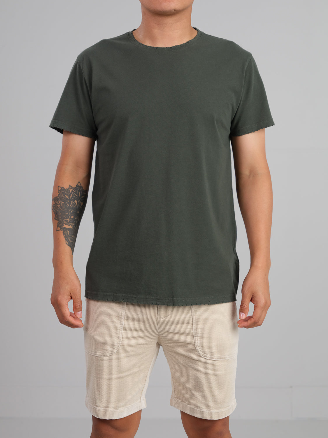A person in a dark green Trekker Tattered Crew Neck Tee by ID Menswear and light beige shorts stands against a plain gray background. The lightweight jersey fabric complements the tattoo on their left forearm.
