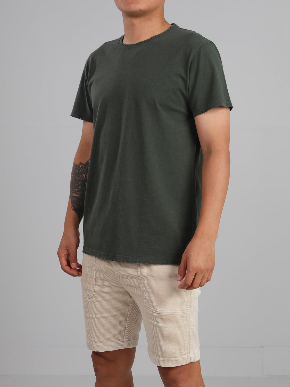 A person wearing an ID Menswear Trekker Tattered Crew Neck Tee in dark green organic cotton, paired with beige textured shorts, poses against a light gray background. The short-sleeve Tee enhances the lightweight jersey feel, and a tattoo adorns their right forearm.