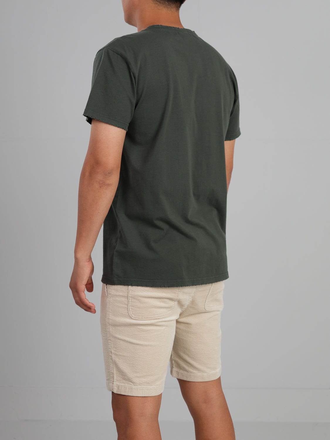 Facing away from the camera, a person wears an ID Menswear Trekker Tattered Crew Neck Tee made of lightweight organic cotton, paired with light beige shorts against a plain gray background.