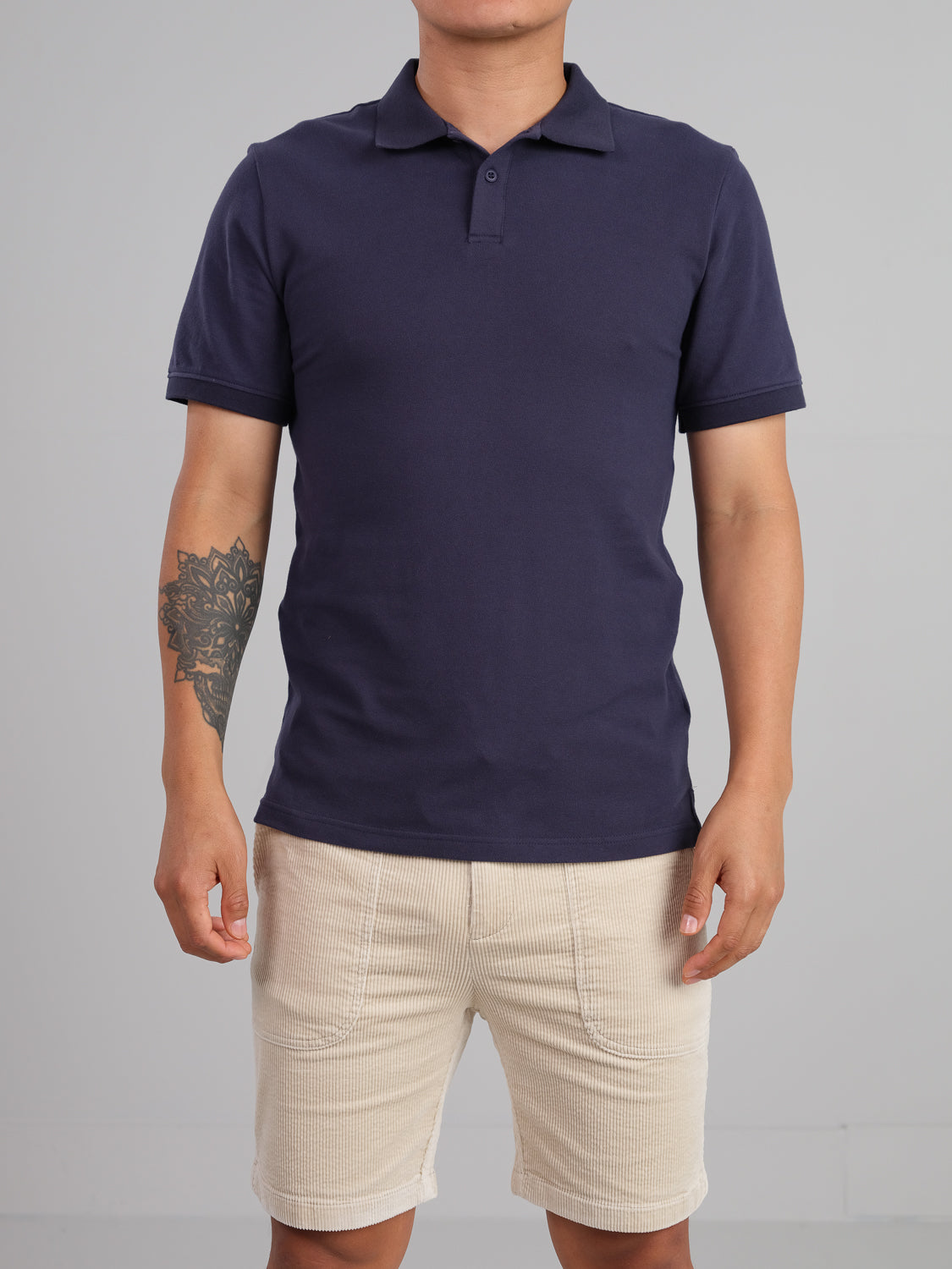 A person sports a Ranger Classic Cotton Mesh Polo by ID Menswear in dark blue and beige shorts against a plain gray backdrop. The lightweight cotton polo has a classic collar and short sleeves, while their left arm displays a geometric tattoo.