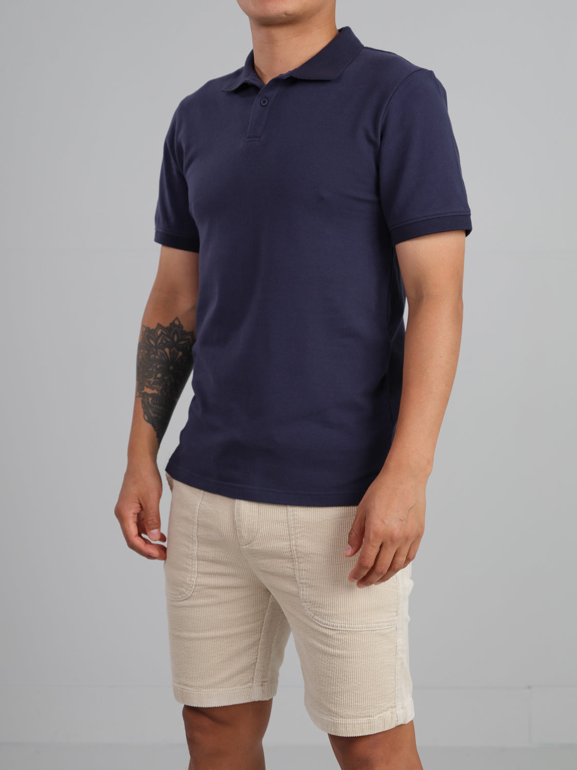 Someone in a Ranger Classic Cotton Mesh Polo by ID Menswear and beige shorts poses against a gray background. The navy, lightweight cotton polo highlights the tattoo on their left arm. Their face remains unseen.