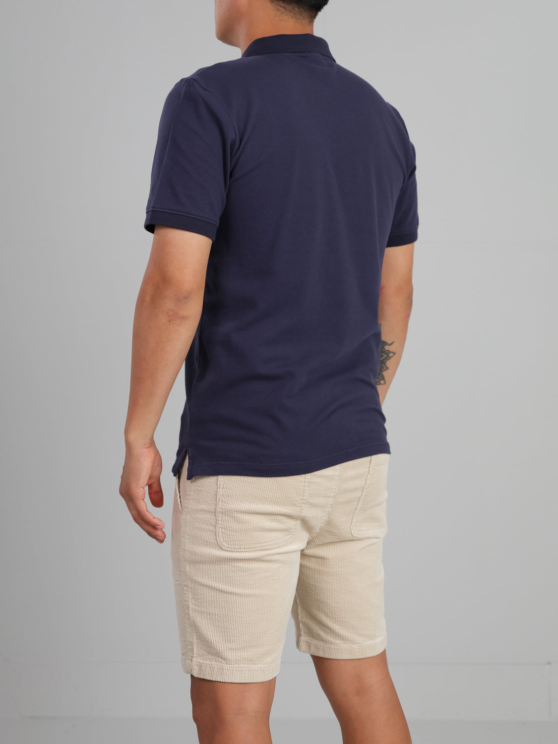 A person, back facing the camera, wears an ID Menswears Ranger Classic Cotton Mesh Polo in dark blue and beige shorts. The lightweight cotton offers a classic style. Relaxed arms expose a small tattoo on the left forearm against a plain backdrop.