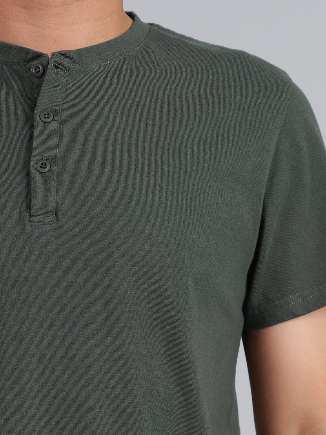 The image highlights the upper body of a person wearing the Champion Organic Cotton Henley by ID Menswear. This dark green, short-sleeve henley made of organic cotton jersey features three buttons and appears on a plain gray background with their head out of frame, radiating understated elegance.