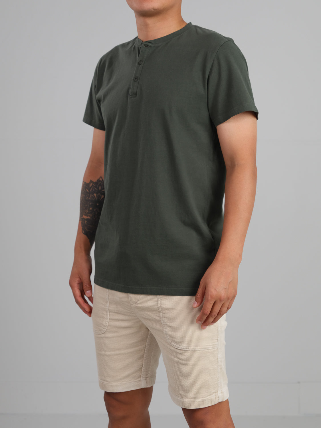 A man stands neutrally, wearing ID Menswears Champion Organic Cotton Henley in dark green and beige shorts, with his left arms tattoo accentuated against a plain gray backdrop.