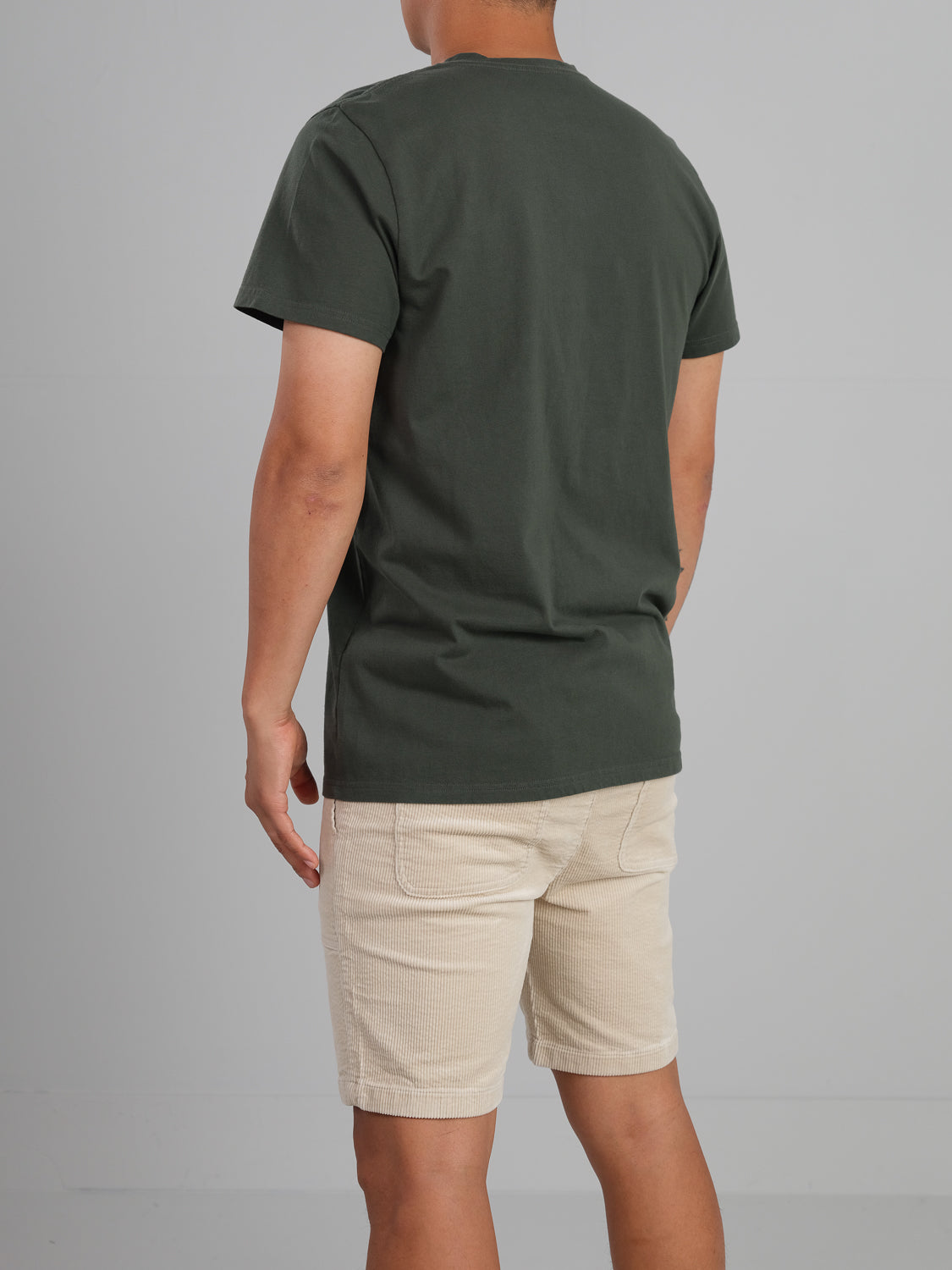 A person faces away from the camera in ID Menswears Champion Organic Cotton Henley, dark green, paired with beige shorts, both crafted from organic cotton jersey, on a plain gray backdrop.