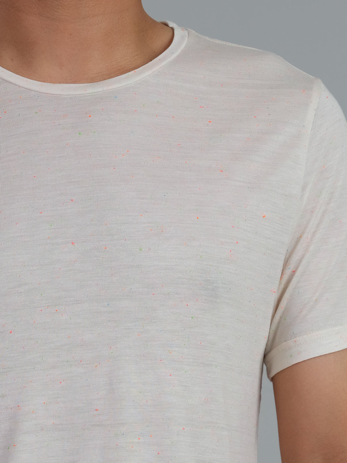 Close-up of someone wearing the Outclass Bamboo Neppy Tee by ID Menswear—a white speckled crew neck T-shirt with tiny colored dots, crafted from lightweight bamboo jersey. The plain gray background complements this design, while the persons face remains out of view.