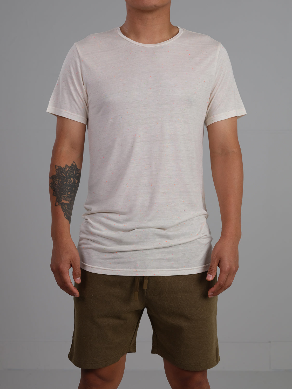 A person wears the Outclass Bamboo Neppy Tee by ID Menswear, paired with olive green shorts. Their right arm showcases a large, intricate black tattoo against a simple gray wall.