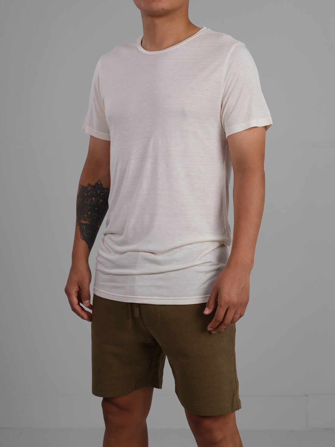 A person wearing the Outclass Bamboo Neppy Tee from ID Menswear, a plain light beige crew neck T-shirt, and olive green shorts stands against a plain gray background, showcasing a tattoo on their right forearm.