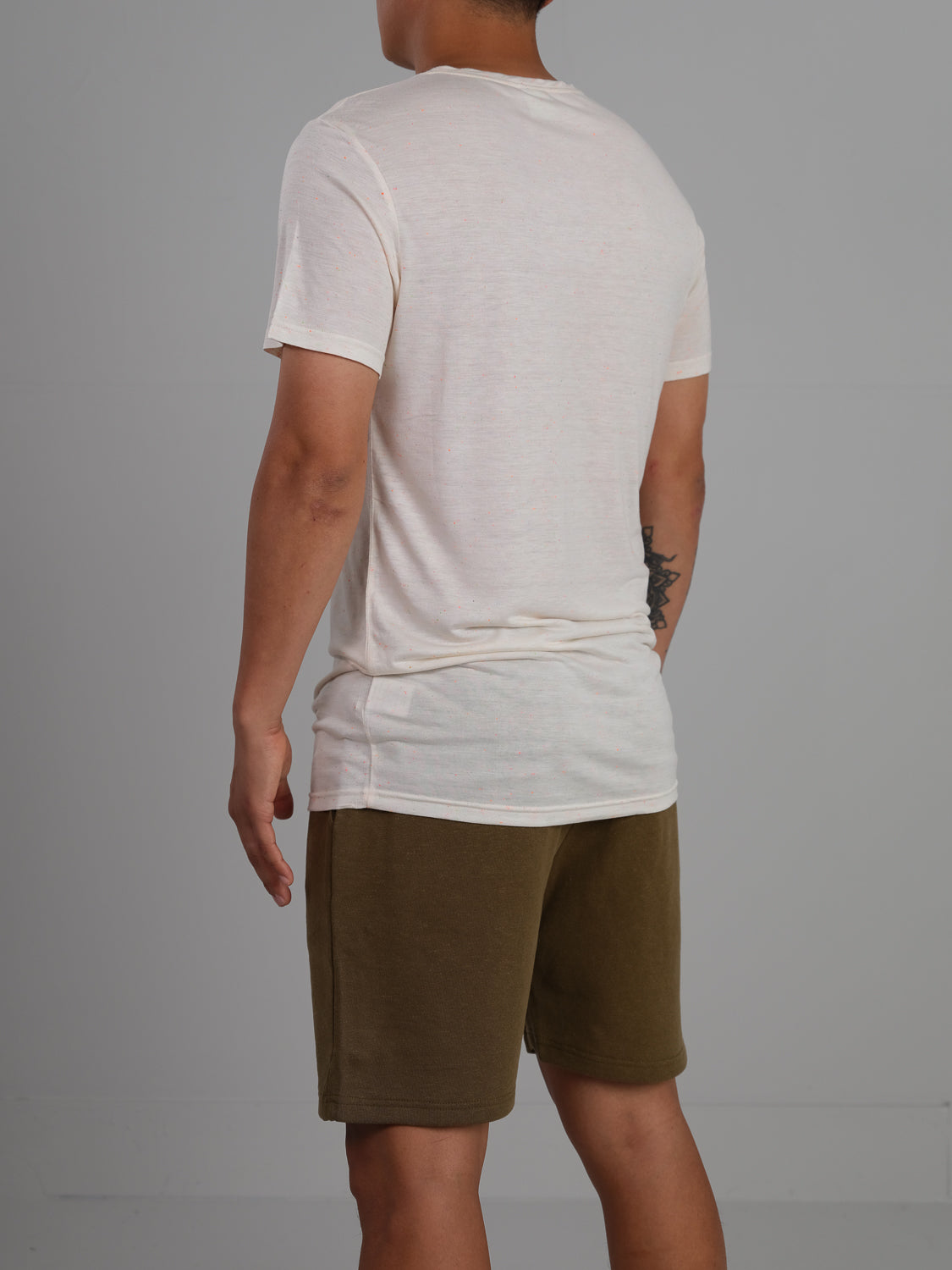 Facing away, a person showcases a tattoo on their right arm while wearing ID Menswears Outclass Bamboo Neppy Tee, a crew neck white t-shirt made from lightweight bamboo jersey for added comfort. They are also in olive green shorts against a neutral gray background.