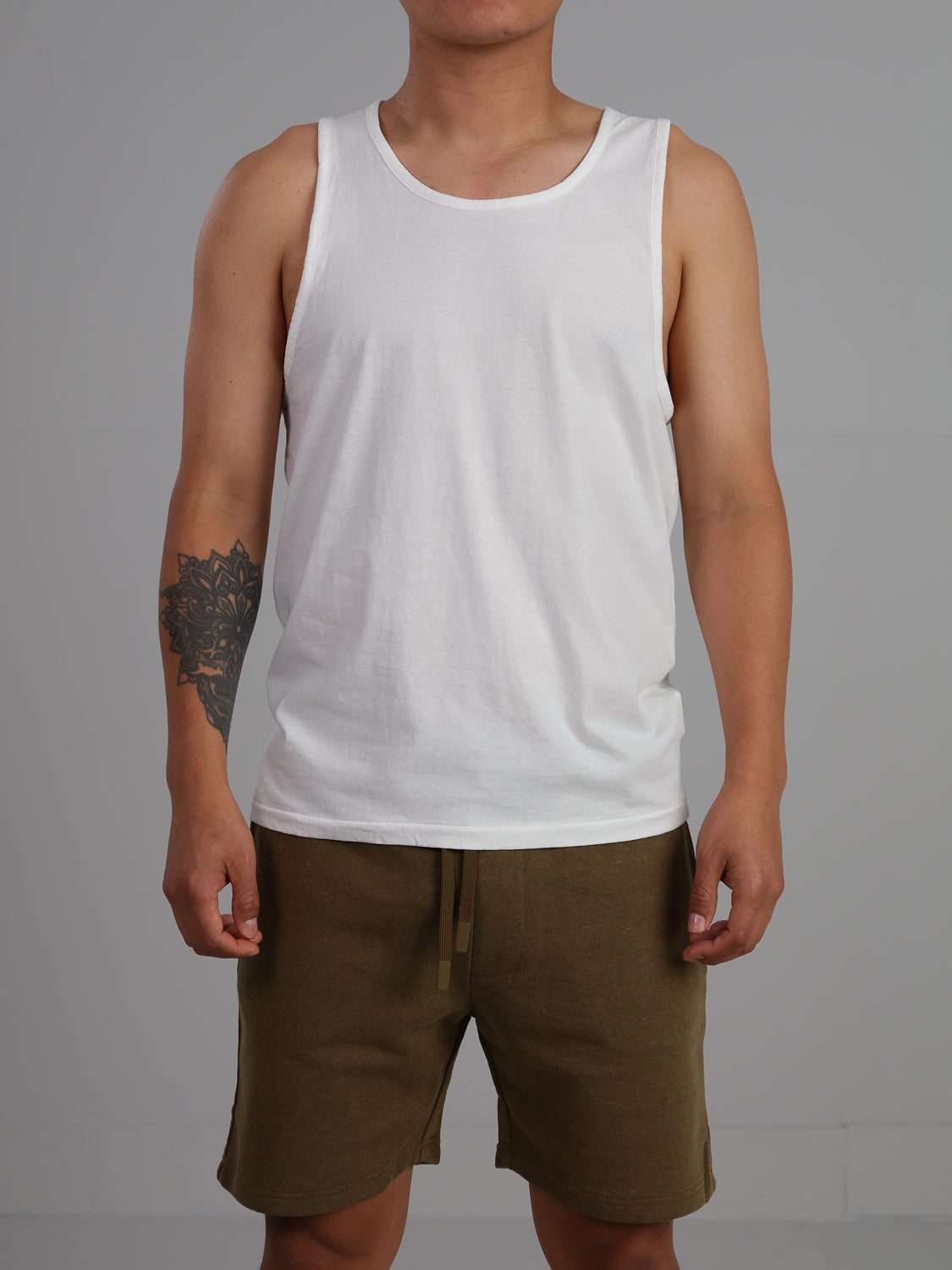 A person models the ID Menswear Settler Organic Cotton Tank Top, known for its lightweight and regular fit, paired with brown shorts. They display a left forearm tattoo against a neutral backdrop. The photo doesnt show their head.
