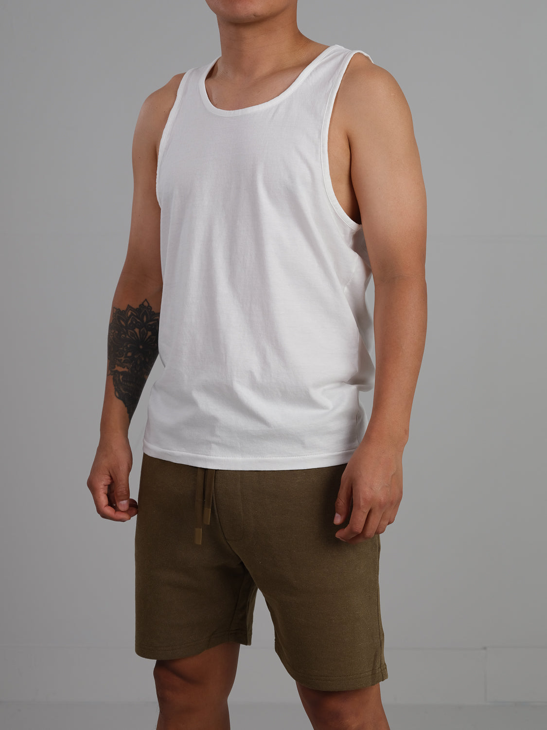 Wearing the ID Menswear Settler Organic Cotton Tank Top and brown shorts, a person stands against a plain gray background. Theres a tattoo on their left arm, and only their upper body and legs are visible.
