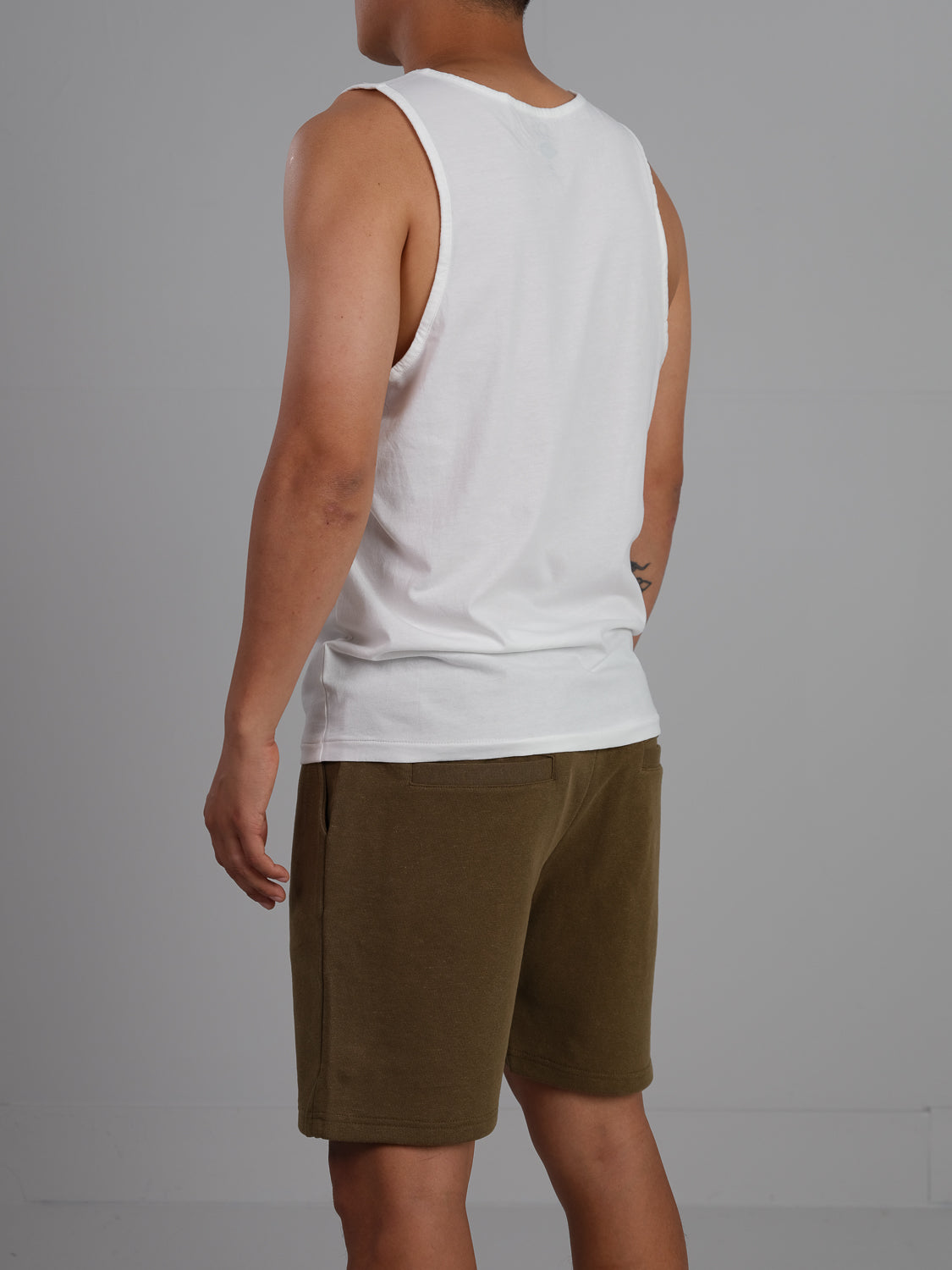 In a gray setting, an individual stands with their back turned, wearing a Settler Organic Cotton Tank Top by ID Menswear and olive green shorts.