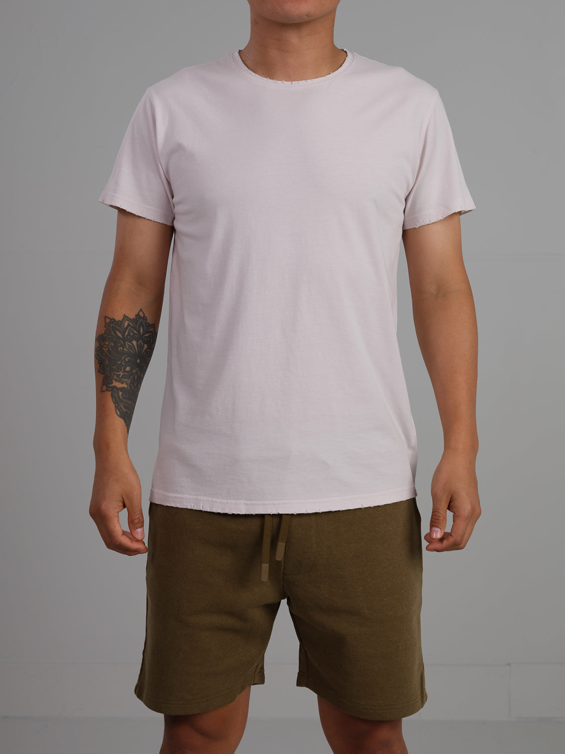 A person in a plain light pink Trekker Tattered Crew Neck Tee by ID Menswear and olive green shorts stands against a gray background, showcasing a tattoo on their left forearm.
