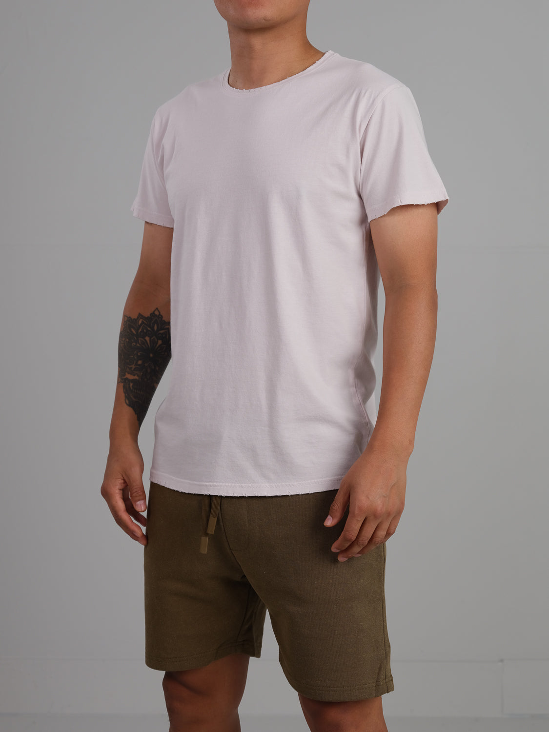 Against a gray background, a person models an ID Menswear Trekker Tattered Crew Neck Tee in plain light pink with olive green shorts, showcasing a forearm tattoo. Their head is out of frame.
