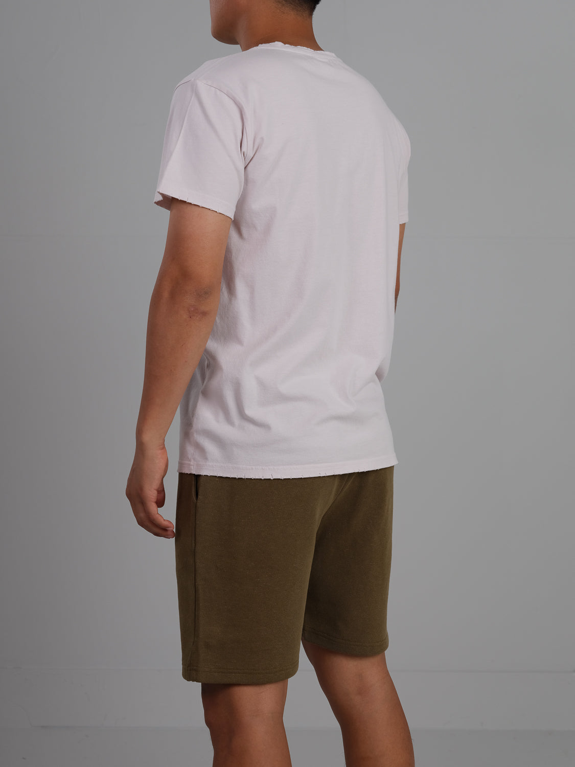 A person in a lightweight ID Menswear Trekker Tattered crew neck tee and olive green shorts stands with their back turned against a plain gray backdrop.